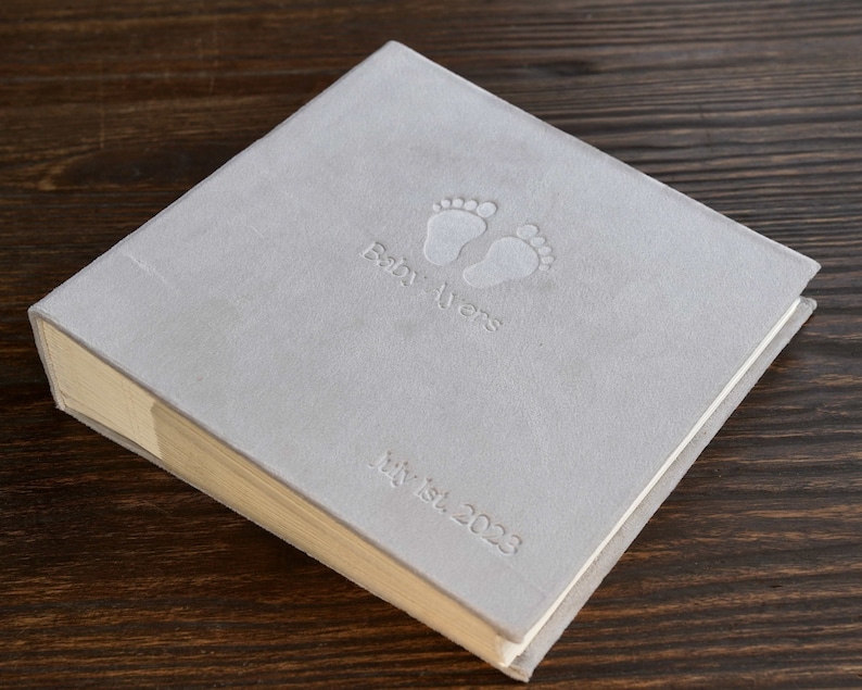 Pode incluir: Um livro de mem&oacute;rias de beb&ecirc; cinza claro com pegadas de beb&ecirc; em relevo e as palavras "Baby Ayers" e "July 1st, 2023". O livro tem uma capa texturizada e uma lombada clara.