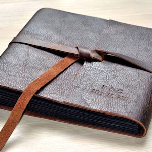 May include: A brown leather photo album with a brown leather strap tied around it. The album is personalized with the initials "F & C" and the date "May 14, 2017".