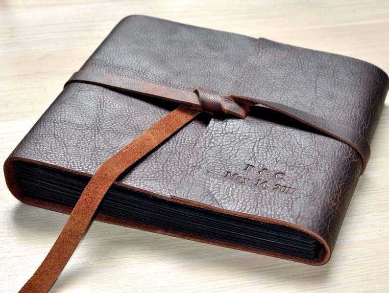 May include: A brown leather photo album with a brown leather strap tied around it. The album is personalized with the initials "F & C" and the date "May 14, 2017".