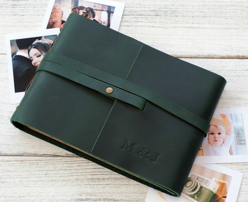 May include: A dark green leather photo album with a brown leather strap and a brass button closure. The album has the initials "M & J" embossed on the front.