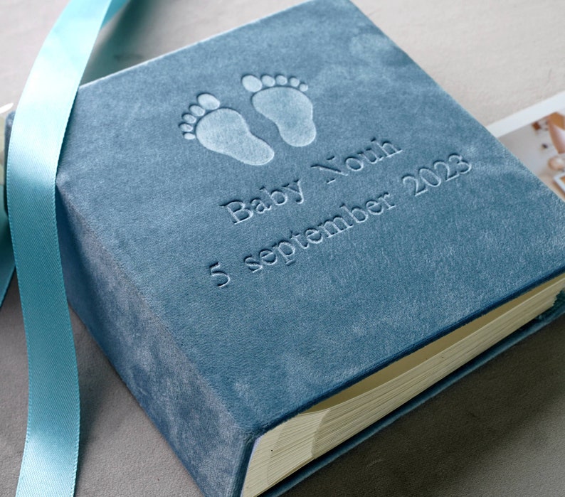 Pode incluir: Um &aacute;lbum de fotos de beb&ecirc; de veludo azul com pegadas de beb&ecirc; em relevo, o nome "Baby Nouh" e a data "5 de setembro de 2023". Uma fita azul clara est&aacute; drapeada sobre o &aacute;lbum. As p&aacute;ginas do &aacute;lbum s&atilde;o vis&iacute;veis.