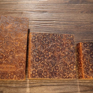 Könnte beinhalten: Drei braune Lederjournale mit einem geprägten Blumenmuster. Das größte Journal hat den Text "Ashley & Justin" und "December 17, 2023" auf der Vorderseite geprägt.