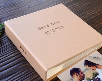May include: A blush-colored photo album with the names "Ben & Anna" and the date "02.12.2025" embossed on the cover. The album is open, revealing a wedding photograph. The album is on a dark wood surface.