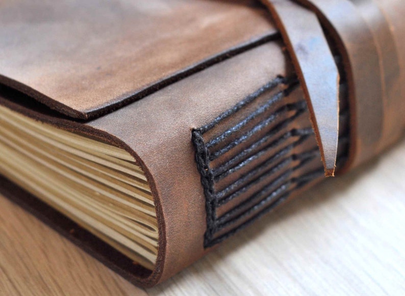 May include: A brown leather-bound journal with black stitching and a leather strap closure. The journal has a rustic and vintage aesthetic.