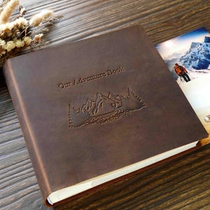 Puede incluir: Un álbum de fotos de cuero marrón con el texto "Our Adventure Book" en relieve en la portada. El álbum presenta un diseño de montañas y árboles.