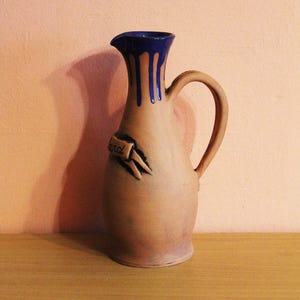 Puede incluir: Una jarra de cerámica con borde y asa azul oscuro. La jarra tiene un cuerpo beige con gotas azules y beige. Una pequeña pancarta con la palabra "and" está adjunta al costado.