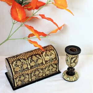 May include: A decorative chest and candlestick holder with a floral design in gold and brown. The chest has a curved top and sits on a black base. The candlestick holder has a black top and base with a gold floral pattern. Orange leaves are in the background.
