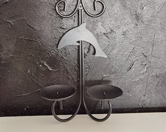 Puede incluir: Un portavelas de metal negro con una silueta de delfín y dos copas para velas. La pieza decorativa tiene una parte superior enrollada y está colocada sobre un fondo gris oscuro texturizado.