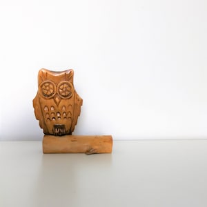 May include: A hand-carved wooden owl figurine with detailed features, including large circular eyes and a textured body. The owl is perched on a small wooden base, showcasing the natural wood grain and color. The figurine is a decorative item.