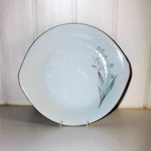 May include: White ceramic plate with a gold rim and a floral design. The plate has a unique shape with a slightly curved edge. The floral design features a cluster of small, light green flowers with blue-green leaves.