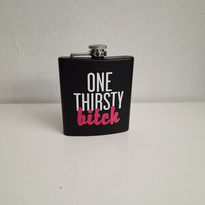 Może przedstawiać: Czarna piersiówka ze srebrną nakrętką. Na piersiówce widnieje napis "ONE THIRSTY bitch" w białych i różowych literach. Piersiówka stoi na białej powierzchni.