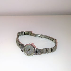 May include: A silver-toned wristwatch with a round face and a linked band. The watch face is gray with red and yellow hands. The band is made of small, rectangular metal links.