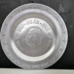 Può includere: Un piatto commemorativo color argento con un bordo floreale inciso. Il piatto presenta un ritratto inciso di un uomo e il testo "1903-80 ÅR-1983" e "OLAV V - NORGES KONGE".
