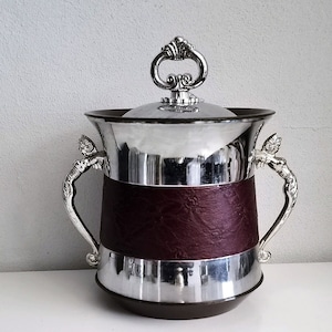 May include: Silver-plated ice bucket with a lid and ornate handles. The bucket features a dark burgundy leather band around the middle. The lid has a decorative handle. A classic piece for serving.