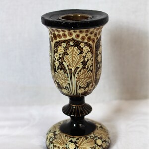 May include: A decorative, ornate candle holder with a black base and rim. The body features a cream-colored background with brown and gold leaf and floral designs. The candle holder is approximately 6 inches tall.