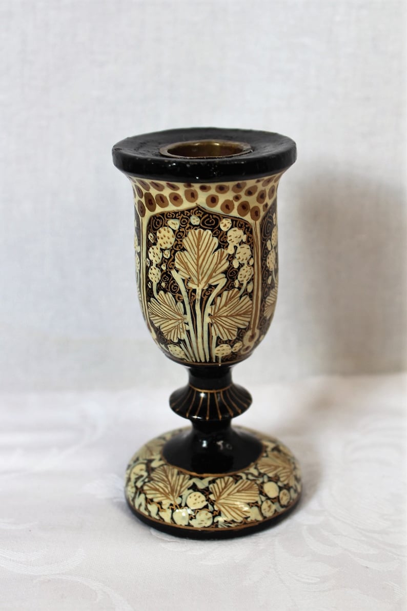 May include: A decorative, ornate candle holder with a black base and rim. The body features a cream-colored background with brown and gold leaf and floral designs. The candle holder is approximately 6 inches tall.