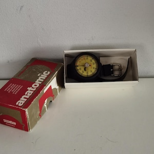 Op de afbeelding: Anatomic duikhorloge met een zwarte band en gele wijzerplaat, gepresenteerd in een witte doos. Het horloge heeft een zwarte lunette en een metalen gesp. Een rood-gouden doos met het woord "anatomic" is ook aanwezig.