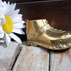 May include: A brass boot-shaped decorative item with a buckle and rivets. The boot is gold-colored and sits on a wooden surface next to white daisies with yellow centers. The background is a dark wooden panel.