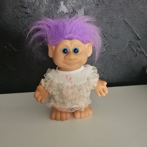 May include: A vintage troll doll with purple hair, blue eyes, and a smiling expression. The doll wears a white dress with a floral pattern and lace trim. The doll is standing on a white surface against a textured black background.