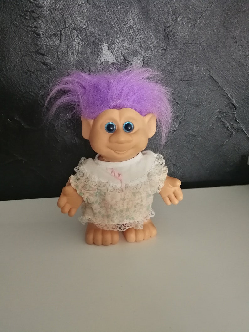May include: A vintage troll doll with purple hair, blue eyes, and a smiling expression. The doll wears a white dress with a floral pattern and lace trim. The doll is standing on a white surface against a textured black background.