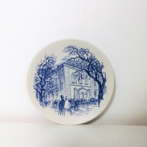 May include: A white ceramic plate with a blue illustration of a building, trees, and people. The plate's design features a detailed architectural drawing, with figures of people in the foreground. The plate is round and has a smooth surface.