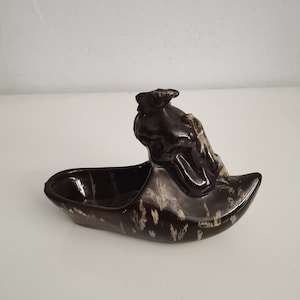 May include: A brown ceramic ashtray in the shape of a boat with a figurehead. The ashtray has a dark brown glaze with lighter brown and beige accents. The boat-shaped ashtray is on a white surface.