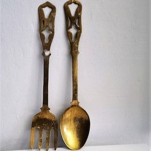 Op de afbeelding: Twee antieke messing serveerbestekken: een vork en een lepel. De vork heeft vier tanden en beide bestekken hebben decoratieve opengewerkte handvatten. Het messing heeft een warme, verouderde patina.