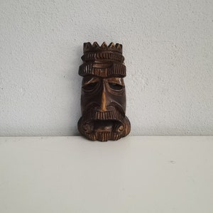Puede incluir: Máscara de madera tallada que representa una cara barbuda con corona. La máscara es marrón y tiene tallas detalladas alrededor de los ojos, la boca y la barba. Probablemente sea un objeto decorativo o una obra de arte.