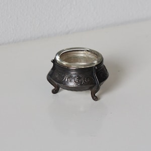 May include: A small, antique silver-toned salt cellar with a clear glass insert. The ornate, footed bowl features decorative scrollwork and a rounded shape. A classic piece for the dining table.