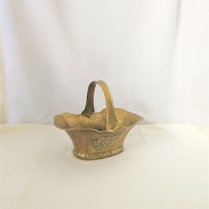 Może przedstawiać: Zabytkowy mosiężny kosz z uchwytem. Kosz ma falisty brzeg i kwiatowy wzór z przodu. Uchwyt jest łukowaty i przymocowany do boków kosza. Kosz jest wykonany z mosiądzu i ma błyszczące wykończenie.