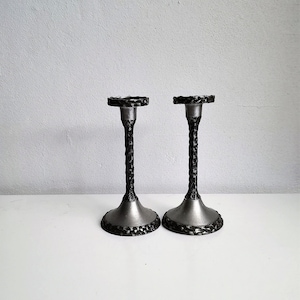 Puede incluir: Dos candelabros de metal plateado con tallos texturizados y bases circulares. Los candelabros tienen un diseño decorativo martillado alrededor de la base y la parte superior. El fondo es una pared gris claro lisa y una superficie blanca.