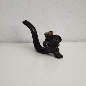 Puede incluir: Una pipa de tabaco artesanal de color marrón oscuro, con una boquilla curvada y una cazoleta esculpida y detallada. El interior de la cazoleta es de color marrón rojizo. La pipa está hecha de madera oscura, posiblemente teñida.