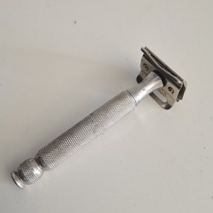 May include: A vintage, silver-colored safety razor with a textured handle. The razor head is rectangular with a blade guard. The handle has a diamond pattern for grip. The razor is designed for shaving.