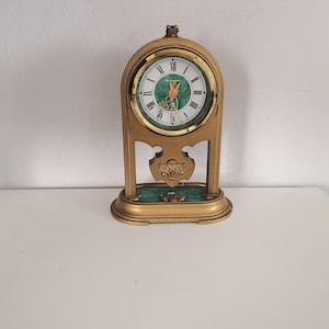 May include: A vintage brass-toned mantel clock with a green and white face. The clock has Roman numerals and decorative accents. The clock is on a white surface.