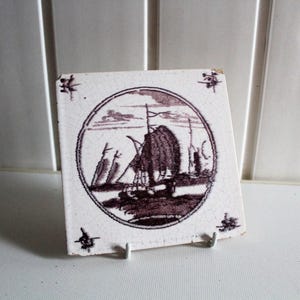 Può includere: Piastrella quadrata in ceramica bianca con un'illustrazione color seppia di una barca a vela in una cornice circolare. La piastrella ha un aspetto leggermente invecchiato ed è sostenuta da un supporto metallico. Elementi decorativi in ogni angolo.