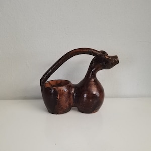 May include: A handcrafted, brown ceramic vase with a unique, organic shape. The vase features a double-lobed base and a curved handle, with a small opening at the top. The vase is approximately 20 cm tall.