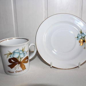 May include: A white porcelain cup and saucer set featuring a gold rim and a painted design of light blue flowers tied with a gold ribbon. The cup displays the text "Voor Oma" in gold lettering, matching the floral motif on the saucer.