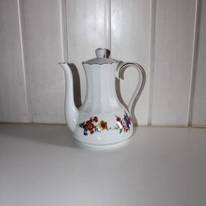 May include: White ceramic coffee pot with a floral design. The pot has a long spout, a curved handle, and a lid. The floral design includes red, yellow, and purple flowers. The pot is approximately 8 inches tall.