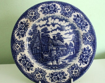 Op de afbeelding: Een ronde, blauw-witte decoratieve plaat met een landelijke scène. De rand van de plaat is versierd met bloemen- en schildmotieven. Het centrale beeld toont een dorpsgezicht met gebouwen en figuren.