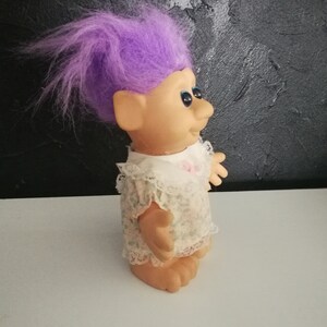 May include: A vintage troll doll with purple hair, blue eyes, and a smiling expression. The doll wears a white dress with a floral pattern and lace trim. The doll is standing on a white surface.