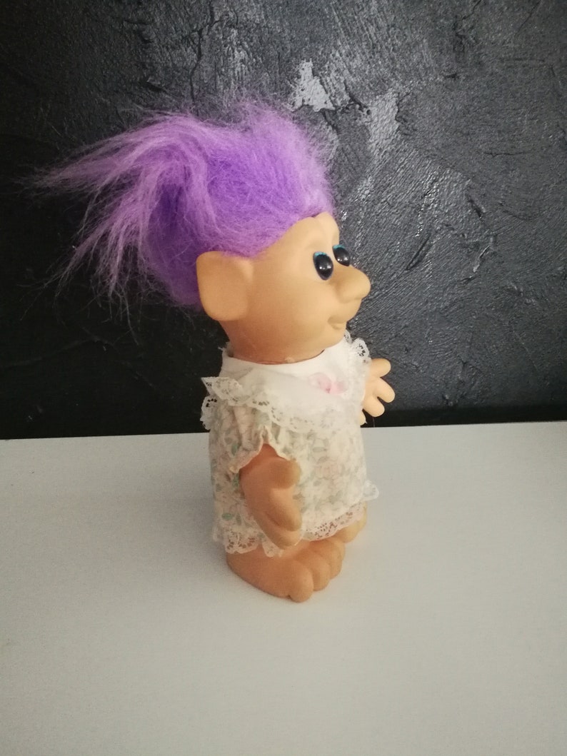 May include: A vintage troll doll with purple hair, blue eyes, and a smiling expression. The doll wears a white dress with a floral pattern and lace trim. The doll is standing on a white surface.