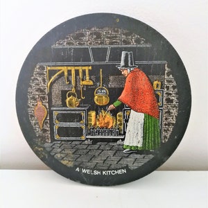 Może przedstawiać: Okrągły, czarny talerz dekoracyjny przedstawiający walijską kuchnię. Kobieta w tradycyjnym stroju zajmuje się ogniem w ceglanym palenisku. Talerz ma złote akcenty i napis "A WELSH KITCHEN".