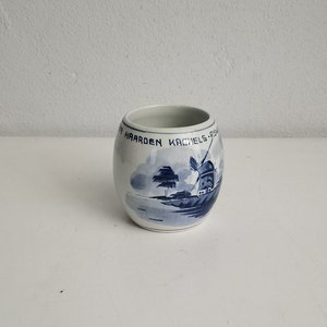 May include: A small, white ceramic cup with a blue and white windmill and landscape design. The cup has text around the rim. The design is reminiscent of Delftware pottery.