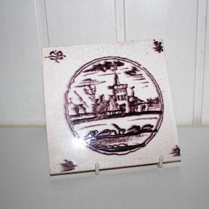 May include: Square ceramic tile with a hand-painted illustration in a circular frame. The image depicts a landscape with buildings and a tower, rendered in shades of purple and white. Decorative floral accents are in each corner.