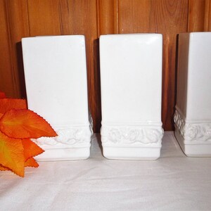 May include: Three white rectangular vases with a decorative floral design at the base. The vases are arranged on a white surface with a wooden background. A cluster of orange and yellow leaves is to the left of the vases.