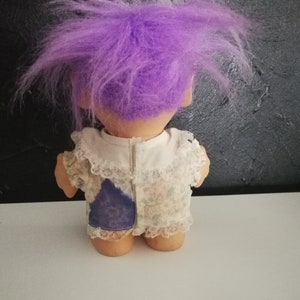 May include: A vintage troll doll with a large, spiky purple hair. The doll is wearing a white dress with a floral pattern and lace trim. A patch of blue fabric is visible on the back of the dress.