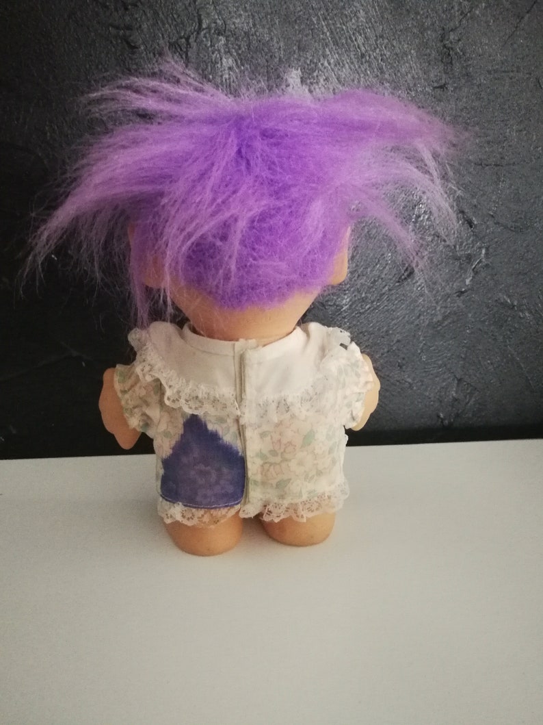 May include: A vintage troll doll with a large, spiky purple hair. The doll is wearing a white dress with a floral pattern and lace trim. A patch of blue fabric is visible on the back of the dress.
