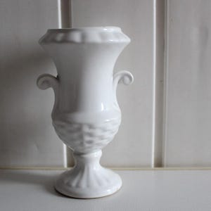 May include: A white ceramic vase with a fluted base and a textured, basketweave-like pattern on the body. The vase has two scrolled handles and a ruffled rim. The background is a white paneled wall.