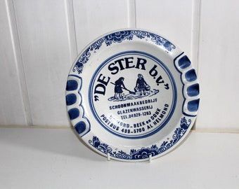 May include: White and blue ceramic plate with a decorative border and text. The plate features a central design with two figures and the text "DE STER b.v.", along with additional text and a phone number.