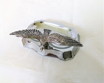 May include: A chrome-plated hood ornament featuring a detailed eagle with outstretched wings. The eagle is silver with textured feather detailing. The base is a stylized shape with a textured section. The ornament is set against a white background.